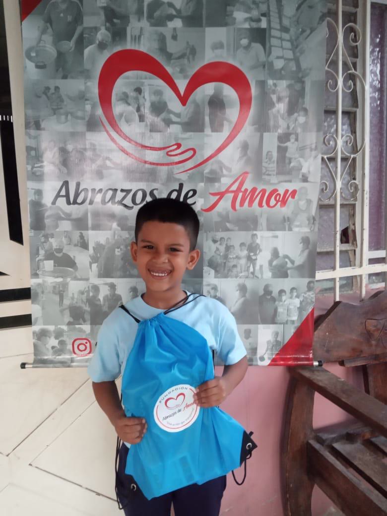 Niño sonriente sostiene su kit escolar frente al mural de la Fundación Abrazos de Amor.